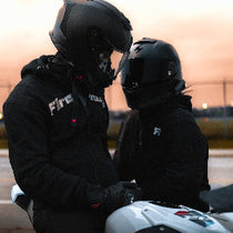 Two people wearing black helmets and jackets on a motorcycle track at sunset.